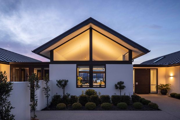 Twilight exterior view of a single-storey contemporary residence arranged around a central gabled pavilion. The home features dark metal pitched roofs, crisp white rendered walls, large black-framed glazing, warm illuminated timber-toned soffits, and carefully clipped ornamental planting along the front entry.