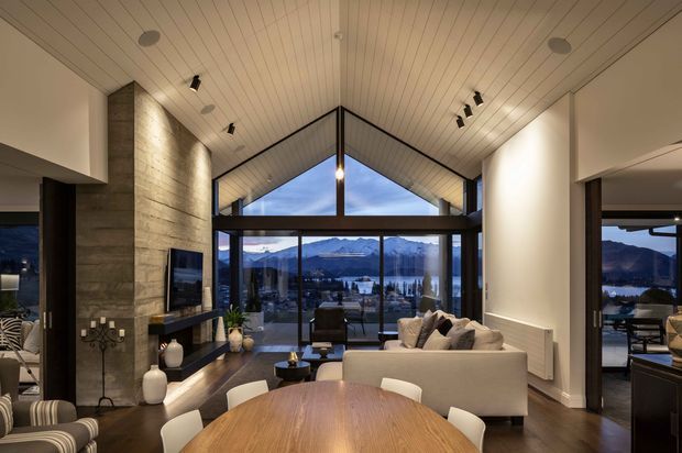 A refined open-plan living and dining area with a dramatic vaulted, timber-lined ceiling and floor-to-ceiling glazing framing a mountain and lake view. The room features a large stone-clad fireplace wall with integrated television, a pale sectional sofa, a round timber dining table, dark timber flooring, and contemporary track lighting.