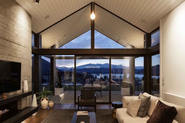 A refined living room with a dramatic vaulted, timber-lined ceiling and exposed dark structural framing. Full-height glazing opens the room to a balcony or terrace with expansive mountain and lakeside views. The interior includes a cream sofa, dark coffee table, television, fireplace feature, and warm ambient pendant lighting.