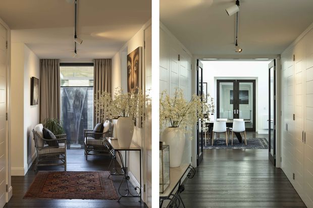 A bright, refined residential hallway and entry sequence with dark timber flooring, white paneled walls and cabinetry, track lighting, large ceramic planters, framed artwork, and woven lounge chairs. The corridor opens toward a glazed front entry and a small seating area with a table and chairs.