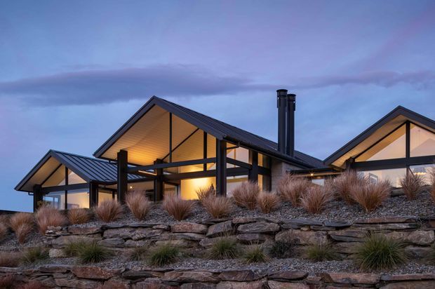 Exterior view of a contemporary multi-gabled home at dusk, with dark standing-seam metal roofs, extensive warm-lit glazing, exposed dark structural posts and beams, and a landscaped terraced rock retaining wall with ornamental grasses in the foreground.