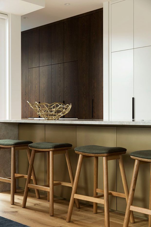 A refined kitchen island with a pale stone benchtop, bronze-toned panelled cabinetry, tall dark timber wall cabinetry, and integrated white cupboards. Several timber bar stools with dark upholstered seats line the island, while a sculptural metallic bowl provides a decorative accent. The space is softly lit and combines warm timber tones with a clean, contemporary built-in design.