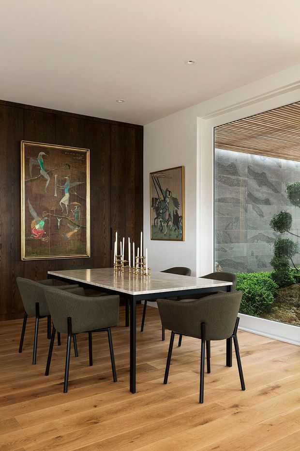 A refined dining room with a rectangular stone-topped dining table surrounded by four upholstered chairs, set against a dark timber-panelled feature wall. Framed artwork, a brass candleholder, recessed ceiling lights, and wide glazing overlooking a landscaped courtyard complete the warm, sophisticated interior.