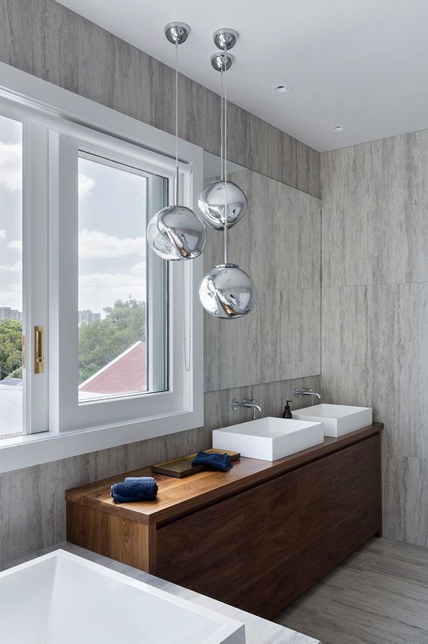 Contemporary bathroom with a double-sink floating timber vanity, white rectangular vessel basins, wall-mounted chrome taps, grey stone-look wall tiling, matching grey floor finish, and a freestanding white bathtub in the foreground. A large white-framed window provides natural light, while three reflective chrome pendant lights hang above the vanity.