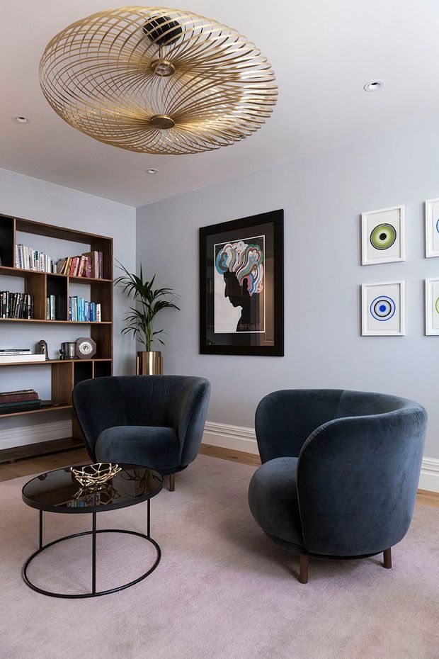 A refined living room with pale grey walls, timber built-in shelving, dark blue upholstered lounge chairs, a black round coffee table, and soft neutral carpeting. A sculptural brass wire pendant light is suspended from the ceiling, complemented by framed contemporary artwork, decorative objects, and a tall indoor plant.