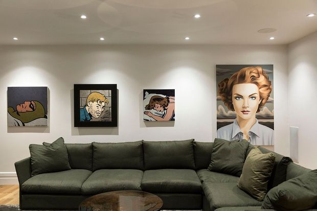 A contemporary living room with a large dark olive-green sectional sofa, recessed ceiling lights, pale walls, and timber flooring. Pop-art portraits and illustrations are arranged across the wall behind the seating, creating a gallery-like focal point. A round dark coffee table is partially visible in the foreground.