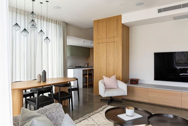 A bright, contemporary open-plan living and dining area. The space features a long timber dining table with black chairs, sculptural pendant lights, sheer floor-to-ceiling curtains, a timber TV and storage wall, white lounge chair, round dark coffee tables, and polished grey concrete flooring. A compact kitchen is visible in the background.