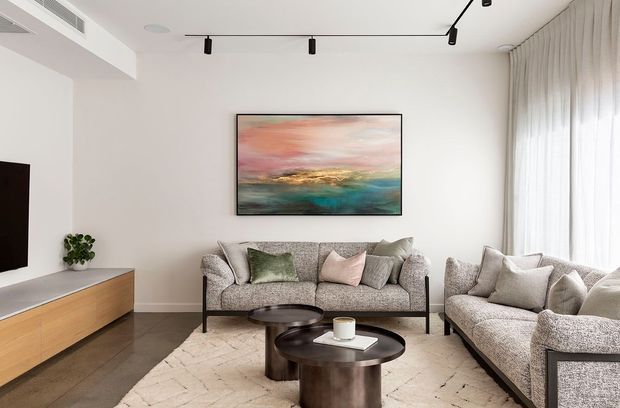A bright, refined contemporary living room with a grey upholstered sofa and matching armchairs arranged around two round dark coffee tables. A large abstract artwork provides a muted pink, teal, and gold accent above the sofa. The room has white walls, full-height sheer curtains, a pale textured rug, dark timber flooring, a wall-mounted television, and a low timber media cabinet. Black track lighting and slim black furniture frames add understated contrast.