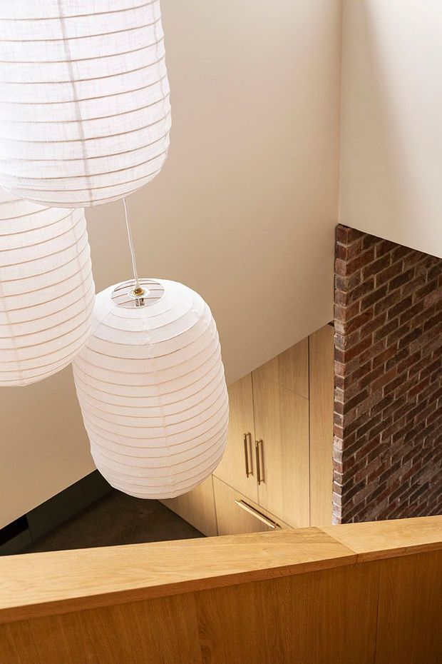 Interior view looking over a timber stair or mezzanine balustrade toward a lower-level space. Three large white paper pendant lanterns hang in a vertical cluster beside warm light-wood cabinetry with slim brass-toned handles. A dark exposed brick wall rises on the right, contrasting with smooth warm beige plaster walls and timber surfaces. The image has a warm, refined contemporary character with a strong vertical composition.
