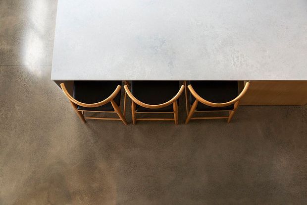 Top-down view of a long kitchen island or counter with three wooden bar stools tucked beneath it. The island has a pale, subtly mottled benchtop and warm timber cabinetry or trim, set against a dark grey polished concrete floor. The composition is minimalist and strongly emphasizes the clean linear arrangement of the counter and seating.