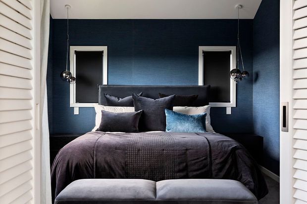 A compact, dark-toned bedroom viewed through partially open white louvred doors. The room features a bed dressed in charcoal bedding with layered grey and blue cushions, a deep navy textured feature wall, two small white-trimmed windows, hanging chrome globe pendant lights, and a grey upholstered bench at the foot of the bed.
