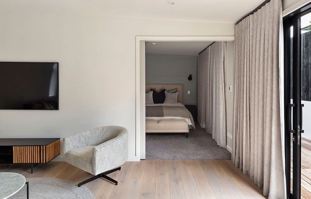 A bright, contemporary living room with pale timber flooring, a wall-mounted television, low timber media console, and a curved upholstered lounge chair. Full-height textured curtains frame black-framed glazing on the right, while an open doorway leads directly into a compact bedroom with a bed and upholstered headboard. The room has a restrained neutral palette and clean-lined detailing.