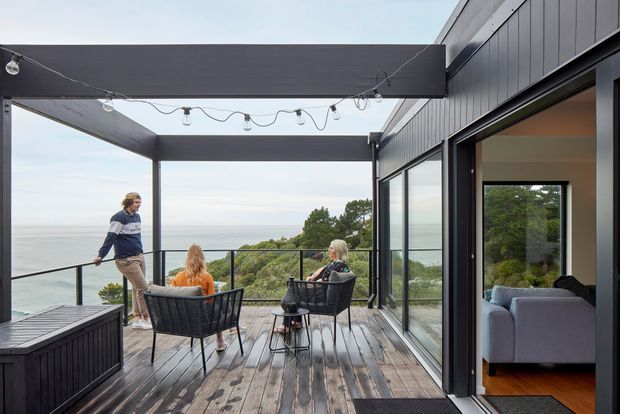A contemporary dark-clad home opens onto a spacious timber deck overlooking a coastal or waterfront landscape. The deck is furnished as an outdoor lounge and dining area beneath a large flat pergola, with exposed string lighting, black metal balustrades, and extensive floor-to-ceiling glazing. A living room with a pale sofa is visible through the open sliding glass doors.