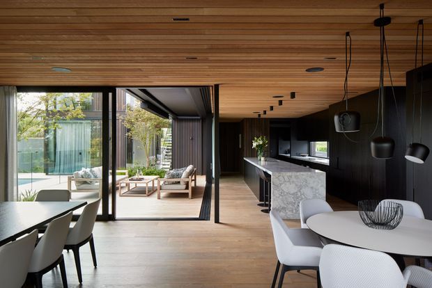 Open-plan contemporary interior combining a dining area, living area and kitchen. The room features extensive timber-lined ceiling and warm timber flooring, dark built-in kitchen cabinetry, a marble-patterned island benchtop, floor-to-ceiling glazing, and multiple black pendant lights. Sliding glass doors open to a landscaped outdoor courtyard or patio.