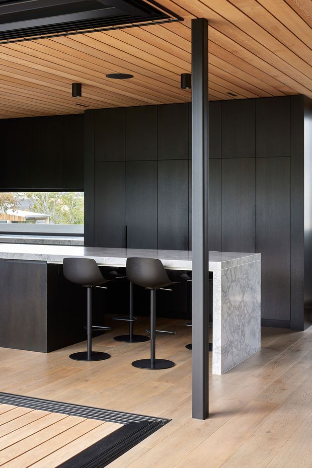 A contemporary kitchen with floor-to-ceiling dark cabinetry, a large pale veined stone island and matching stone benchtops, black bar stools, and warm timber flooring and ceiling lining. A slender dark structural post divides the view, while expansive glazing brings in daylight and views of trees outside.