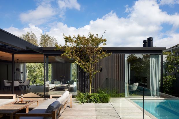 Contemporary single-storey courtyard residence with dark vertical cladding, extensive floor-to-ceiling glazing, and a covered outdoor entertaining area opening onto a tiled pool terrace. The image shows a timber outdoor deck, built-in or integrated seating, dining furniture, landscaped planting, and a linear swimming pool beneath a flat roofline.
