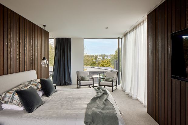 A refined bedroom with a large upholstered bed, dark accent cushions, and a textured throw. Vertical dark timber wall panelling wraps the room, complemented by soft carpet, full-height sheer curtains, and a small seating area beside expansive glazing overlooking trees. The room has a calm, warm contemporary character with strong indoor-outdoor connection.