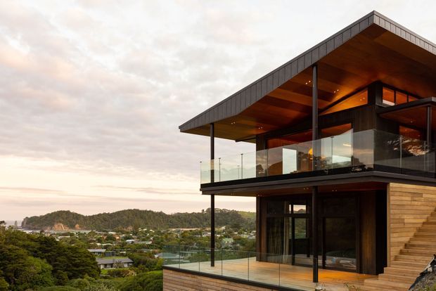 Exterior architectural photograph of a contemporary multi-level hillside home with a dramatic projecting flat roof, dark structural framing, extensive floor-to-ceiling glazing, and glass-balustraded balconies. Warm timber soffits and timber-clad walls contrast with the dark facade. The elevated terrace overlooks a green suburban valley and wooded hills.