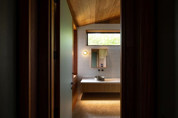 A compact contemporary bathroom viewed through a doorway, featuring a floating timber vanity with a vessel basin, a rectangular wall mirror, small round wall light, fine light-coloured wall tiles, a timber-lined sloping ceiling, and a narrow horizontal window. Warm concealed lighting beneath the vanity and natural timber surfaces create an intimate, refined atmosphere.