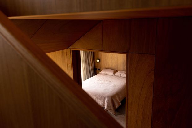 A warmly lit view through angular timber-lined framing into a compact bedroom. The room contains a bed with pale bedding, bedside lighting, and curtains, while the foreground is dominated by rich dark wood panelling and geometric built-in elements.