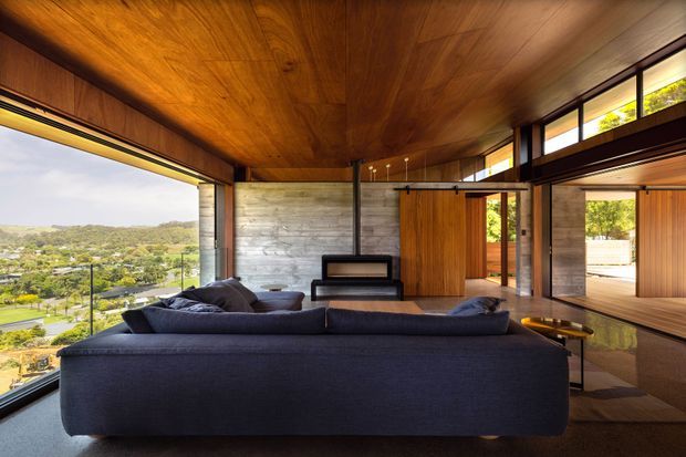 A contemporary living room with a large dark sectional sofa, timber-lined ceiling and walls, exposed concrete feature wall, and expansive floor-to-ceiling glazing overlooking a green suburban landscape. Sliding timber doors and black-framed glazing connect to an adjacent terrace or outdoor space, while a linear pendant light and minimalist fireplace reinforce the refined modern design.