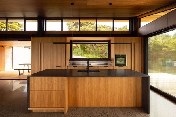 A warm, contemporary kitchen viewed from the living area, featuring a large central island with dark stone benchtop, timber-grain cabinetry, integrated sink and tap, and built-in oven. Floor-to-ceiling glazing and high clerestory windows bring in views of dense greenery, while timber-lined ceilings and dark window frames create a strong connection to the surrounding landscape.