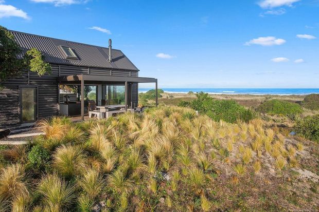 Exterior view of a dark-clad coastal house set among native dune grasses and scrub, with a covered outdoor living area and expansive ocean horizon beyond. The house has a steep pitched metal roof, dormer window, chimney, timber screening, and large glazed openings. Bright natural daylight and an informal seaside landscape create a relaxed coastal atmosphere.