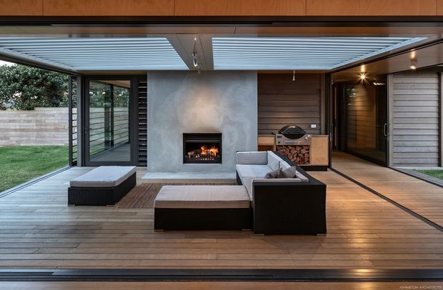 A covered, open-sided outdoor living area with a timber deck, dark-framed glazing, a central concrete fireplace, built-in wood storage, barbecue, and low outdoor lounge furniture. Adjustable louvred roof panels provide filtered daylight, while timber wall cladding and warm integrated lighting create a sheltered, contemporary entertaining space connected to a lawn and garden.