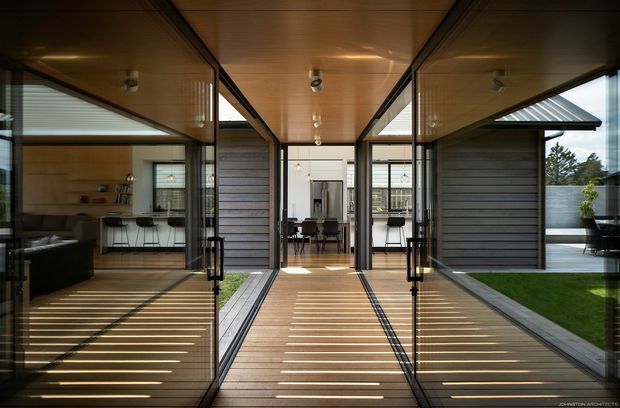 A covered central passageway or glazed link between wings of a contemporary home. The corridor features warm timber ceiling and decking, extensive black-framed glazing, dark horizontal-clad walls, and views into a kitchen on the left and dining area ahead. A small landscaped courtyard with lawn and concrete fencing is visible to the right.
