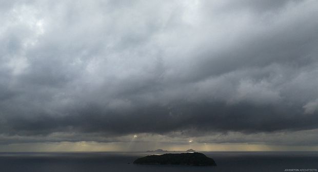 A moody seascape showing a small dark island or rocky headland surrounded by calm water beneath dramatic, heavy storm clouds. The image is predominantly atmospheric and landscape-focused, with no visible building or interior space.
