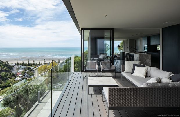 A contemporary high-level coastal apartment terrace overlooking a beachfront and ocean. The covered balcony features dark timber-look decking, frameless glass balustrades, an outdoor lounge setting with a woven sectional sofa and armchairs, and a marble-look coffee table. Full-height dark-framed glazing opens into a refined open-plan living and kitchen area with dark cabinetry and stone surfaces.