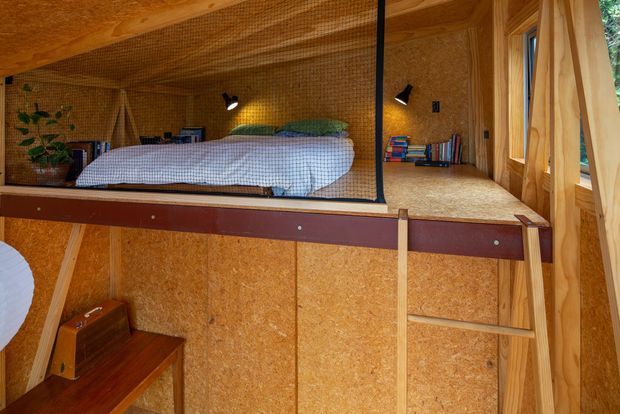 Compact loft-style sleeping nook enclosed in warm wood-toned oriented strand board, with a double bed, built-in bedside shelves, wall-mounted reading lights, timber ladder access, and a small wooden desk or bench below. The space has an intimate, cabin-like appearance and visible structural timber framing.