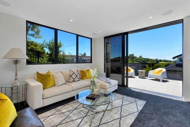 Bright contemporary living room with a cream sectional sofa, yellow accent cushions, glass coffee table, patterned area rug, and large black-framed corner glazing. The open sliding doors connect directly to a sunny terrace with outdoor seating and views of surrounding greenery.