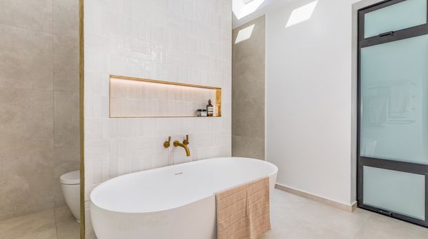 Bright contemporary bathroom featuring a freestanding white oval bathtub, wall-mounted brass tapware, white textured wall tiles, a recessed display niche with warm integrated lighting, and large-format beige stone-look wall and floor finishes. A partially concealed toilet and dark-framed frosted-glass door are visible. Skylights provide strong natural illumination.