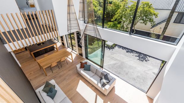 High-angle view of a bright contemporary double-height living and dining space. A timber dining table sits beside a slatted timber balustrade and built-in feature fireplace, while sofas occupy the lower level. Floor-to-ceiling glazing, a large glazed opening, suspended sculptural pendant lights, and a courtyard or roof terrace visible beyond create an airy, open interior.