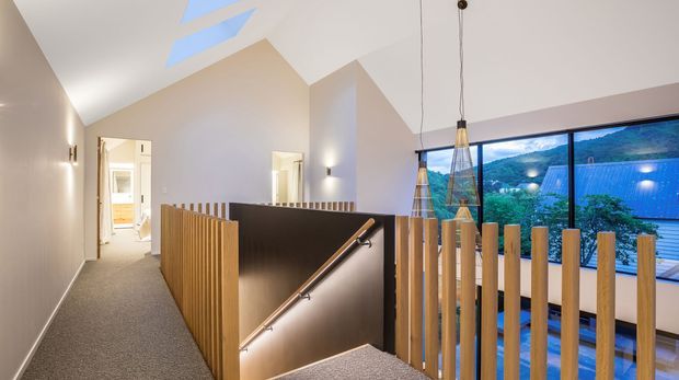 A bright contemporary upper-level hallway and stair landing with a stairwell enclosed by vertical timber balustrades and a dark feature wall. The space has a carpeted floor, white and pale neutral walls, angular vaulted ceiling forms with skylights, slim wall lighting, and pendant lights. Large floor-to-ceiling glazing overlooks a green mountainous landscape and neighbouring rooftops.