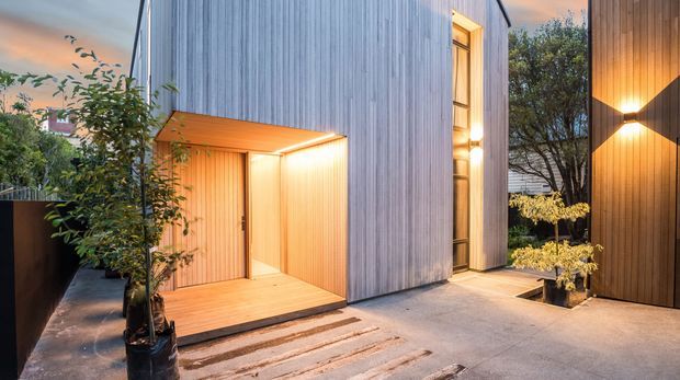 Contemporary architectural exterior courtyard at dusk, featuring vertically clad timber volumes, a warmly lit recessed entry porch, tall narrow windows, concrete paving, and landscaped planters. The composition highlights the building facade and sheltered outdoor arrival space.