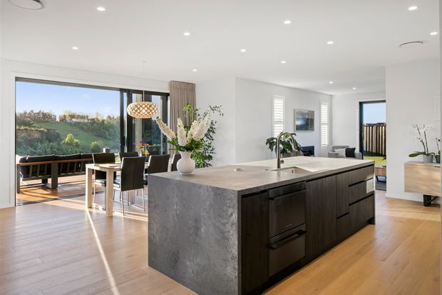 Bright open-plan kitchen and dining area with a large central island featuring a grey stone waterfall benchtop and dark timber cabinetry. The room has light timber flooring, white walls and ceiling with recessed downlights, expansive black-framed glazing opening to an outdoor deck, and a dining table beside the kitchen. Contemporary, spacious, and airy interior.