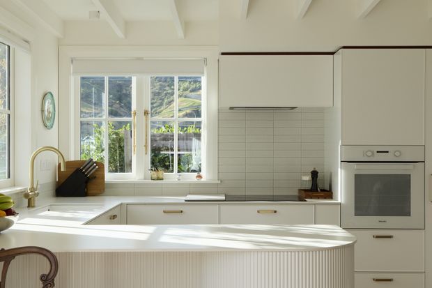 Bright, compact kitchen with a curved island featuring vertical fluted detailing, white shaker-style cabinetry, a pale horizontal-tile splashback, integrated oven, and a farmhouse-style sink beneath multi-pane windows. Exposed white ceiling beams and warm timber hardware add traditional character, while strong natural light creates an airy atmosphere.