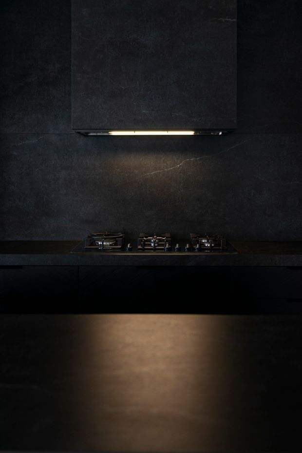 A dramatic close-up of a dark, minimalist kitchen cooking zone. Three gas burners are set into a black benchtop beneath a large dark rangehood, with matching dark wall cladding or splashback and black cabinetry below. A warm strip light under the rangehood provides the main illumination, creating a moody, refined atmosphere.