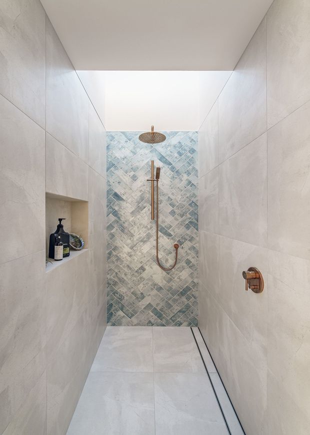 A minimalist walk-in shower with large pale stone-look wall and floor tiles, a blue-grey patterned feature tile wall, a rainfall showerhead, handheld shower, and warm copper-toned plumbing fixtures. A recessed wall niche holds toiletries. The space is bright, clean, and contemporary with a skylight or overhead opening.