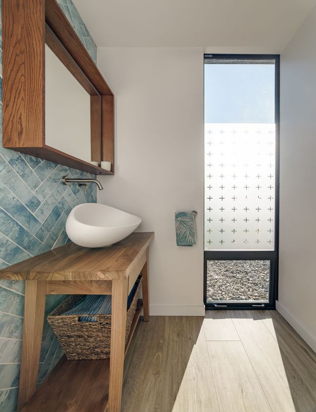 A bright, narrow contemporary bathroom featuring a white vessel basin on a substantial timber vanity, a large timber-framed wall mirror, and blue-green patterned wall tiles arranged diagonally. A tall black-framed window or glazed door with a translucent patterned screen brings in strong natural light. The floor combines warm wood-look flooring with a light tile area near the glazing.