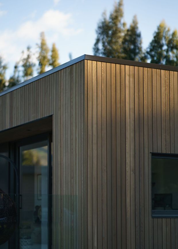 Close-up exterior architectural photograph of a contemporary, box-like building with vertical timber cladding, dark metal roof edge and window frames, and large glazed openings. The simple flat-roofed facade is lit by warm low-angle sunlight, with blurred trees and sky in the background.