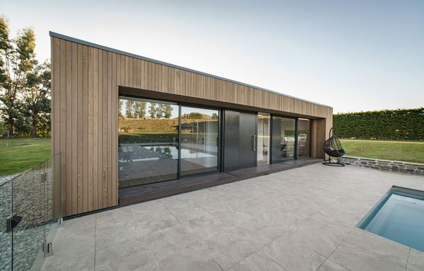 A sleek single-storey modern pavilion or extension beside a swimming pool. The building has a long, low rectangular form with vertical timber cladding, a dark flat roof edge, extensive floor-to-ceiling glazing, and dark-framed sliding doors. It opens onto a large pale stone-paved terrace with frameless glass balustrading, manicured hedges and lawn beyond. The interior is visible through the glazing but is secondary to the exterior architecture.