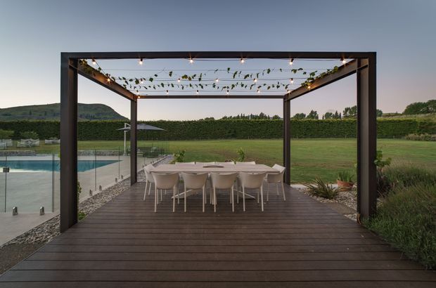 A landscaped outdoor dining terrace with a dark timber deck and a large open pergola structure. The pergola is decorated with festoon string lights and climbing greenery, and shelters a long outdoor dining table with pale chairs. A swimming pool is visible to the left, with expansive lawn, hedges, ornamental planting, and rolling terrain beyond.