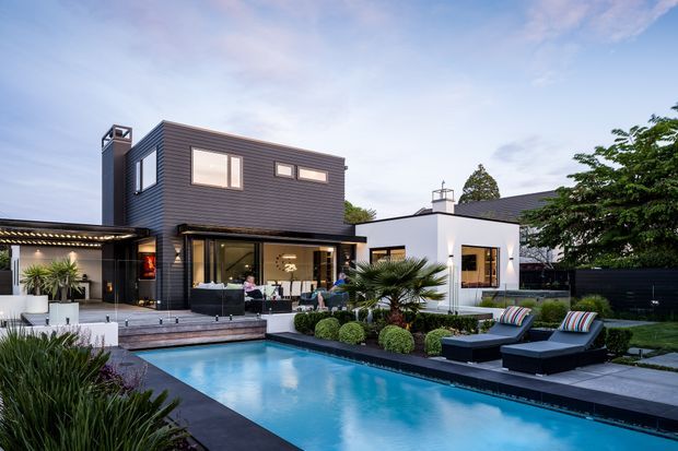 Exterior view of a contemporary multi-level residence with dark horizontal cladding, white rendered sections, expansive glazing, and a landscaped backyard pool terrace. The pool is framed by paved decking, clipped shrubs, palms, outdoor loungers, and a covered outdoor entertaining area.