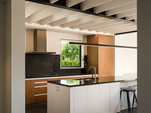A contemporary kitchen with a large central island, integrated sink and chrome tap, dark stone benchtops and splashback, white island cabinetry, and warm timber cabinetry along the rear wall. The room features exposed white ceiling beams, black structural accents, a window overlooking greenery, and streamlined bar seating.