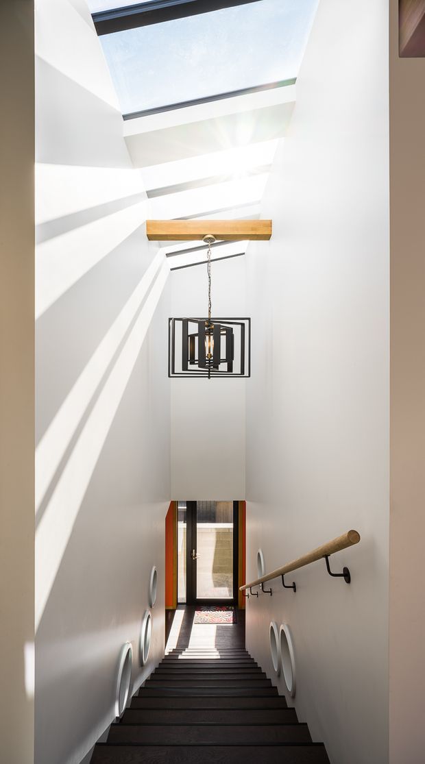 A dramatic narrow stairway descending through a tall white stairwell. Multiple skylights flood the space with sunlight, creating strong linear shadows. A timber beam spans the stairwell beneath a modern black geometric pendant light, while dark stair treads, a timber handrail, black brackets, and circular wall details add contrast. A glazed doorway is visible at the lower landing.