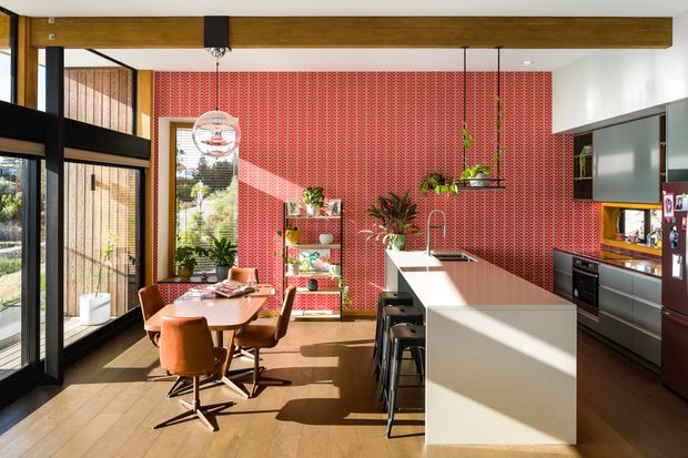 A bright, open-plan kitchen and dining area with a long white island, dark cabinetry, integrated appliances, and a small dining table with rust-coloured chairs. The room features expansive black-framed glazing and doors, warm timber flooring, a substantial exposed timber beam, and a bold red patterned feature wall. Indoor plants, pendant lighting, and strong sunlight add warmth and character.