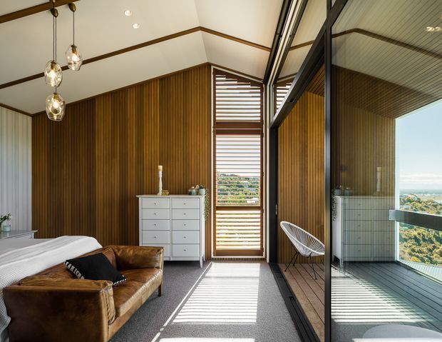 A bright, contemporary bedroom with a pitched ceiling, warm vertical timber wall panelling, carpeted floor, and a large glazed sliding door opening to a balcony or terrace. The room includes a bed, leather lounge chair, white drawer unit, pendant lights, and a louvred timber screen creating strong linear shadows. A scenic coastal landscape is visible through the glazing and reflected in the glass.